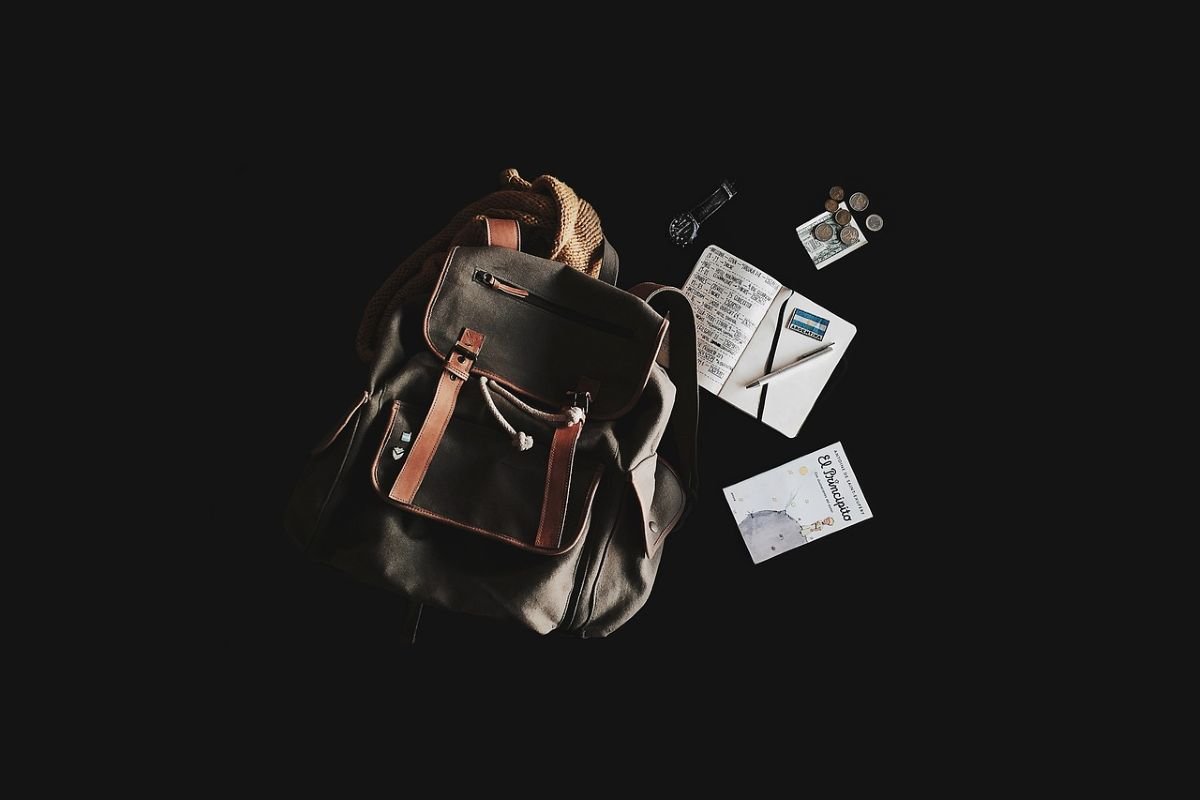 A dark green backpack with brown leather straps, accompanied by a notebook, pen, coins, and a travel guide.