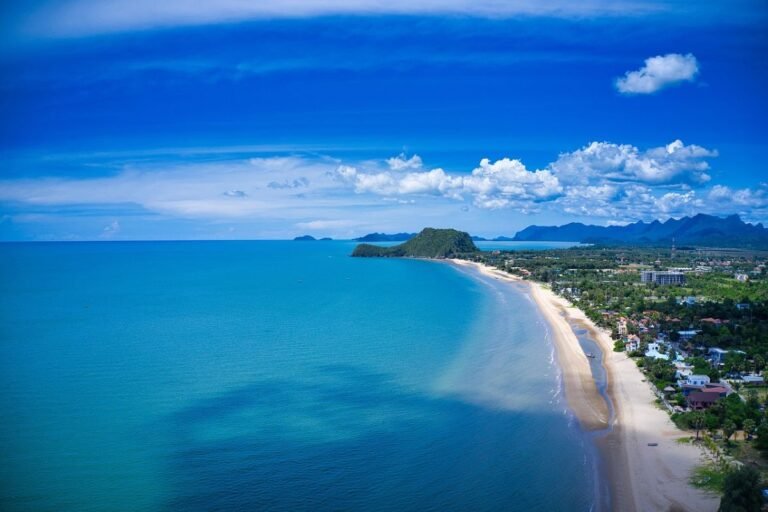 Aerial view of a coastline featuring a sandy beach, clear blue water, and distant mountains under a partly cloudy sky.